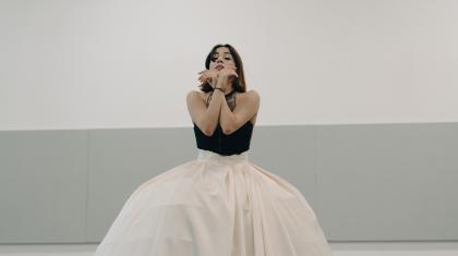 Dancer wearing a voluminous white skirt holding her forearms crossed in front of her face.