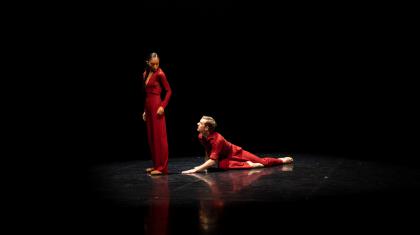 Two dancers dress in red, one is on the floor looking up at the other who is turned away but sees him over her shoulder
