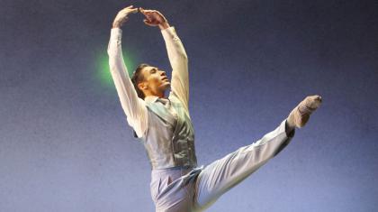 Male dancer stood against a blue stage with a green light behind him. His arms are raised and one leg is extended.