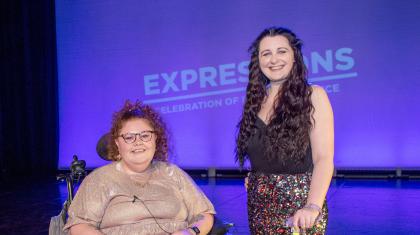 Two presenters are on stage at expressions. The woman on the left is in a wheelchair wearing a sparkly gold dress and red hair. The woman on the right is stood up with a walking stick, wearing a black top, gold sequin skirt and long black hair.