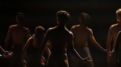 Several dancers, each looking away from the camera, hold the same pose in darkness - their left arm hanging loose and their right to the side, palm up, as if holding something weighty.