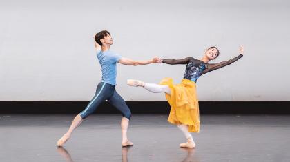 Two smiling dancers, one supporting the other with an outstretched hand, and the other, in a yellow dress and dark patterned top, stands on one leg, her other stretched out in front of her