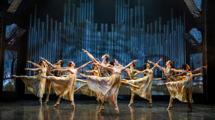 A dozen dancers in gold and white dance in unison, each stood on one foot, the other leg in arabesque behind them