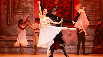 Paris dressed in a masque and in black provides Juliet with balance by holding her raised arm as she stands en pointe with a leg extended behind her