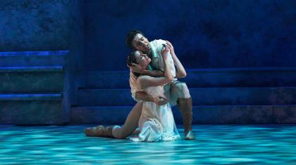 Romeo embraces Juliet as then kneel on the floor, bathed in a cool blue light