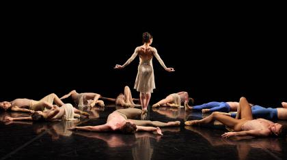 A group of dancers lie on the ground, apparently unconscious, while another stands and walks through the fallen bodies
