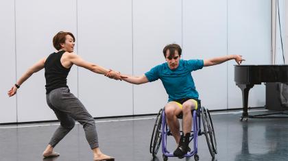 Two dancers hold hands and pull away from each other, one braced with his feet on the ground, the other with the wheels of his chair against the pull