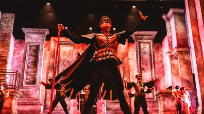 Male dancer wearing a masque on his face, his legs spread firmly apart, holding a red staff in his hand, one end on the floor