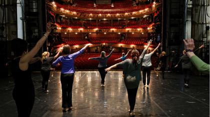 A group of older dancers on a stage stand separately and rehearse dance moves towards an empty multi-tiered audience space