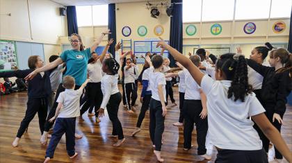 A group of children in a school hall, all barefoot, striking the same dance pose as each other - stretching one arm up and forward, the other down behind them as they step forward, their weight on the front foot