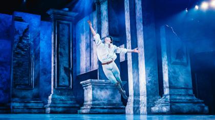 Dancer leaping across the stage, his legs together, one arm raise triumphantly above him, bathed in blue light
