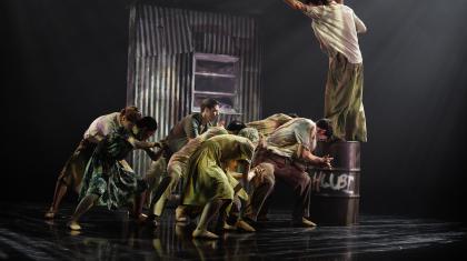 A group are each leaning low, as if burdened, another stands on a metal drum shouting at them