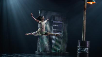 Dancer wearing a light dress leaps gracefully high above the stage, the set of a ramshackle building behind her