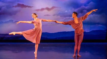 A male and female dancer hold hands. The female dancer lifts her leg and reaches to the left, as the male dancer floats his arm upwards and smiles at her