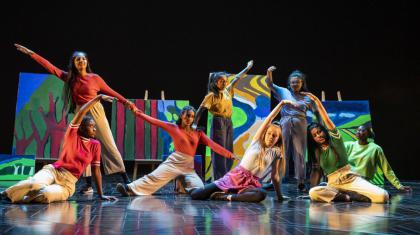 Performers wearing different colours pose on a colourful stage. Some are sat and some are standing and all reach their arm outwards in different direction.