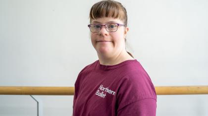 Woman with light brown hair, glasses, and a plum T-shirt with the logo of the Academy of Northern Ballet