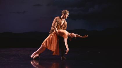 Dancer in an orange dress leans back, she is held by a man in brown with his arms around her waist