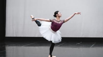 A dancer stood on one leg with the other raised and arm extended. The dancer is wearing a dark purple leotard, black leggings and a white tutu