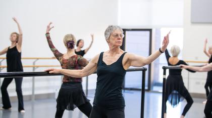 Class of dancers at the barre, one hand holding on for balance, the other arm held out, palm up forearm raised, knees slightly bent