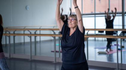 Student in glasses and dark T-shirt, arms raise above her head, participating in a class for people over 55