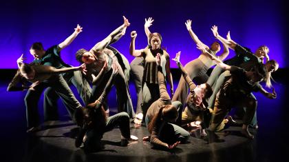 Against a blue and purple backdrop, a figure stands with one arm raised in a defiant gesture surrounded by dancers shielding themselves from her brilliance.