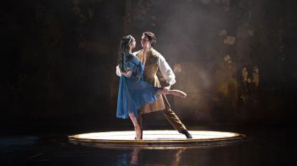 In an illuminated circle two people dance, a woman en pointe in a blue dress and a man who his holding her lovingly
