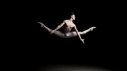 Male dancer, only wearing white leggings, mid jump, legs split, arms reaching down