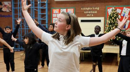 Young girl with straight shoulder-length hair stands in a school hall with her class-mates, arms wide, hands raise and smiling