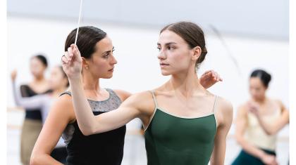 Dancer with feather in hand looks over her shoulder at another placing a hand on her shoulder attentively