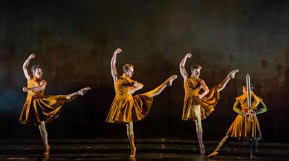 Three men in golden uniforms standing on one foot, their other leg raised, straight at the knee and one arm aggressively above their head, a fourth man is stood facing in a different direction wearing the same uniform but holding a sword behind his back