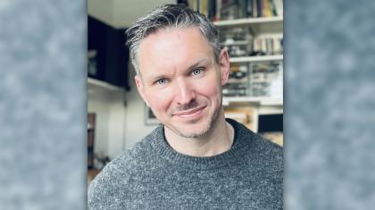 Person presenting male, grey eyes, blue/grey knitted jumper, salt-and-pepper hair, smiling warmly
