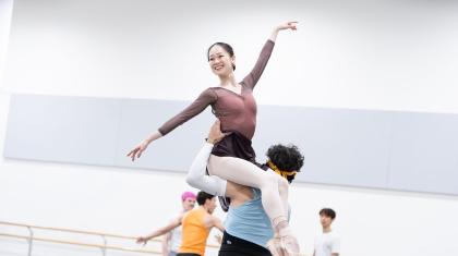 Dancer smiling as she is lifted, sat on the left shoulder of another dancer, one arm angled up, the other angled low