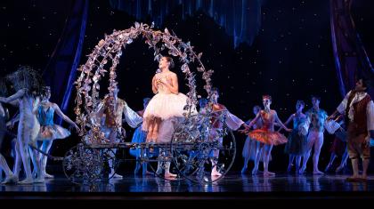 Dressing white, a dancer rides an open, decorated carriage surrounded by onlookers