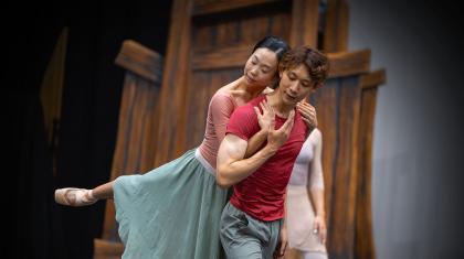 Dancer in a red t-shirt move forward, another holds him from behind and is lifted as she hooks her arms under his and holds his shoulders