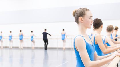 Students in blue leotards stand at the barre, holding it for balance as they strengthen their arches.