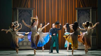 In a dressing room a group of women wearing similar dresses with different accent colours dance around Anne Lister who lifts her hat in respect to them