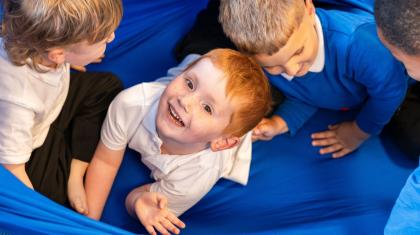 Three young school boys play on top of a bright blue fabric. The boy in the middle looks up at the camera with a smile.