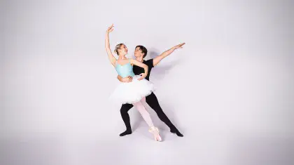 Two student dancers against a pale pink backdrop, one in a blue leotard and tutu on pointe and leaning supported by the other all in black with an arm around her waist