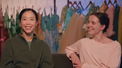 Two people sat on a sofa smiling with costumes hung on a rail in the background