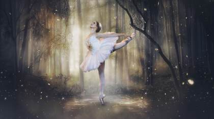 In the middle of a dark forest with soft sunlight, a dancer in a white feathery tutu poses in an attitude, with her back arched and her arms reaching to the back.