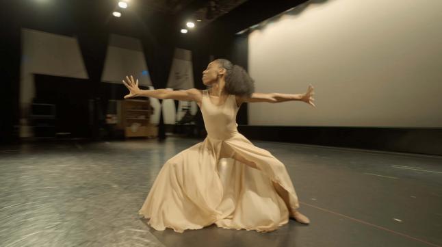 Female dancer in a pale beige dress performs on an empty stage, her arms out to either side of her, her legs bent, and looking to her right